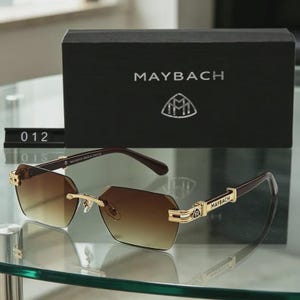 May include: Brown gradient lens sunglasses with gold detailing and the brand name "MAYBACH" on the arms. The sunglasses are presented with a black box displaying the brand logo and the name "MAYBACH".