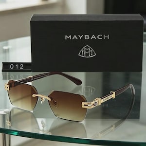 May include: Brown gradient lens sunglasses with gold detailing and the brand name "MAYBACH" on the arms. The sunglasses are presented with a black box displaying the brand logo and the name "MAYBACH".