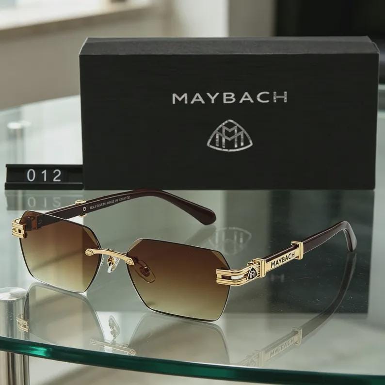 May include: Brown gradient lens sunglasses with gold detailing and the brand name "MAYBACH" on the arms. The sunglasses are presented with a black box displaying the brand logo and the name "MAYBACH".