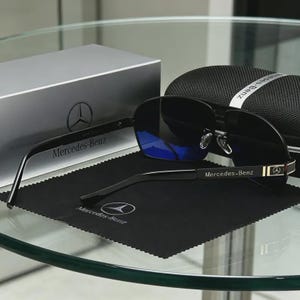 May include: Black sunglasses with blue lenses, a black case, and a silver box, all bearing the Mercedes-Benz logo. The sunglasses rest on a black cleaning cloth. The glasses have a gold accent on the temples.