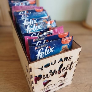 May include: A wooden cat food organizer with the words "YOU ARE Purrfect" cut out on the front. The organizer is filled with blue and pink Purina Felix cat food packets.
