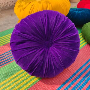 May include: A round, plush purple velvet cushion with a central button and radial pleats. Other cushions in yellow, red, green, and blue are visible in the background. The cushions are on a colorful striped mat.