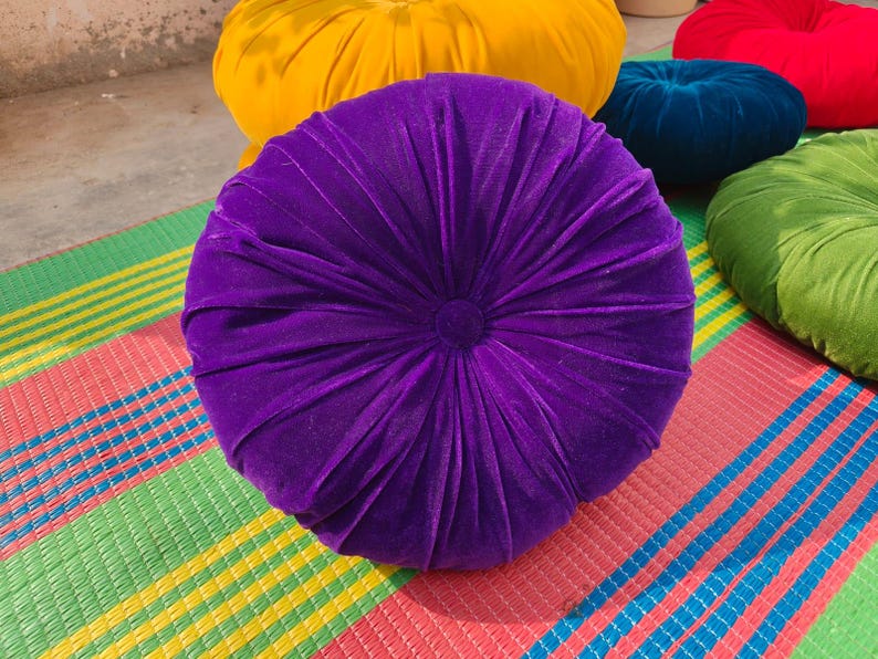 May include: A round, plush purple velvet cushion with a central button and radial pleats. Other cushions in yellow, red, green, and blue are visible in the background. The cushions are on a colorful striped mat.