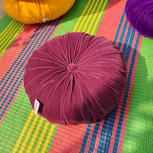 May include: A burgundy velvet round cushion with a central button detail, resting on a colorful striped mat. Other cushions in yellow and purple are also visible. The mat has green, red, blue, and yellow stripes.