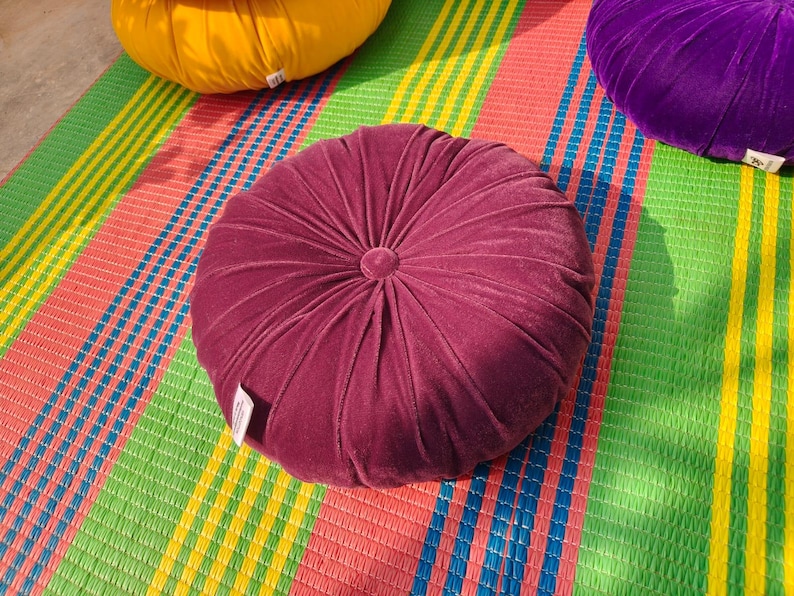 May include: A burgundy velvet round cushion with a central button detail, resting on a colorful striped mat. Other cushions in yellow and purple are also visible. The mat has green, red, blue, and yellow stripes.