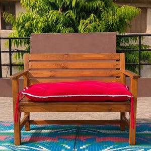 May include: A wooden bench with a slatted backrest and armrests, featuring a rectangular red cushion with white piping trim and side ties. The bench is constructed from light brown wood with a visible grain, providing a sturdy seating structure.