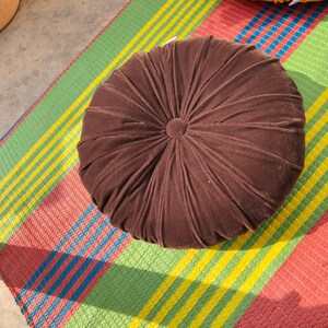 May include: A round, brown velvet cushion with a central button detail. The cushion is placed on a colorful striped mat with red, green, yellow, and blue stripes. The cushion's design features radial stitching.
