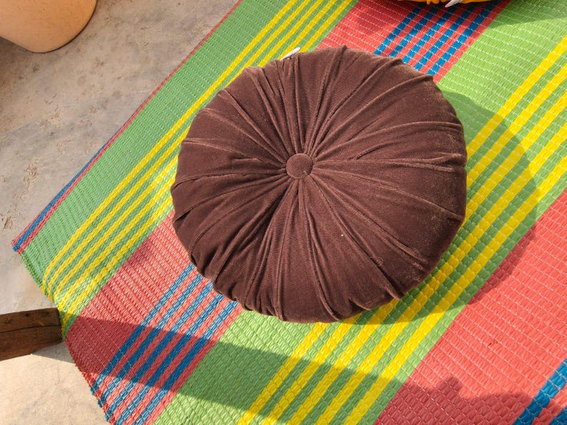 May include: A round, brown velvet cushion with a central button detail. The cushion is placed on a colorful striped mat with red, green, yellow, and blue stripes. The cushion's design features radial stitching.