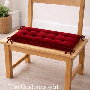 May include: A wooden chair with a rectangular, tufted, burgundy cushion tied to the seat with burgundy ribbons. The text "The Cushions edit" is at the bottom of the image. The chair is made of light-colored wood.