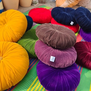 May include: A collection of round, velvet throw pillows in various colors, including yellow, red, green, navy blue, brown, purple, and burgundy. The pillows have a buttoned center and are arranged on a striped mat.