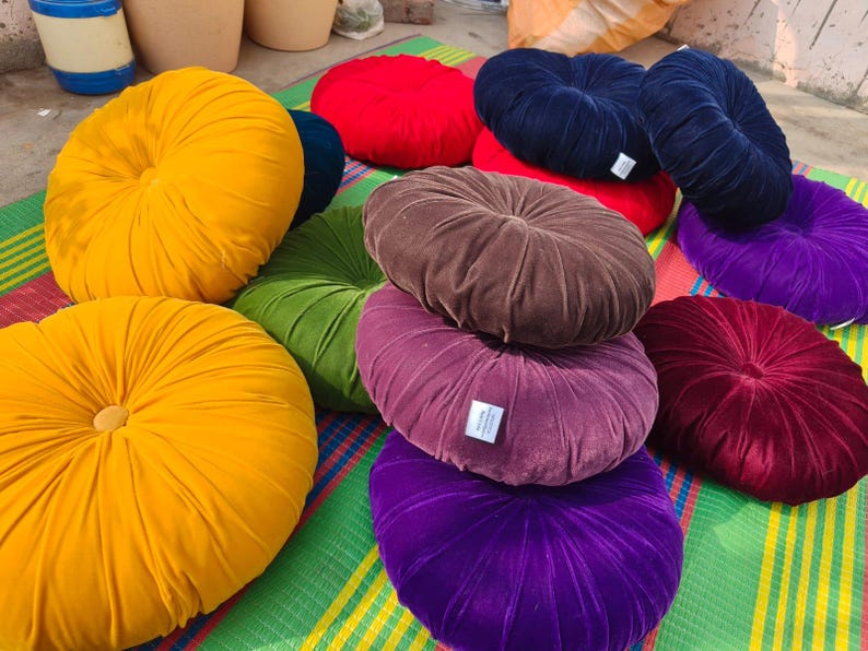 May include: A collection of round, velvet throw pillows in various colors, including yellow, red, green, navy blue, brown, purple, and burgundy. The pillows have a buttoned center and are arranged on a striped mat.