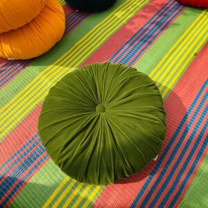 May include: A round, olive green velvet cushion with a button center. The cushion is on a colorful striped mat with red, yellow, green, and blue stripes. Other round cushions in orange, red, and dark green are also visible.