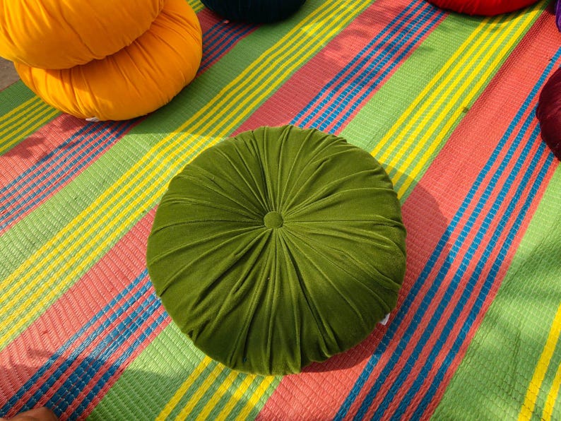 May include: A round, olive green velvet cushion with a button center. The cushion is on a colorful striped mat with red, yellow, green, and blue stripes. Other round cushions in orange, red, and dark green are also visible.