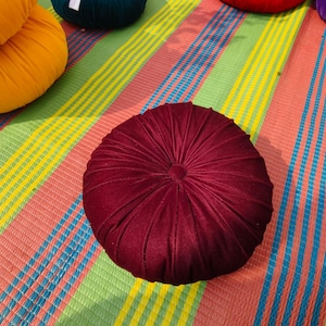May include: A round, burgundy velvet cushion with a central button and radial stitching. The cushion is on a colorful striped mat with shades of pink, green, yellow, and blue. Other cushions in yellow, green, and red are visible in the background.