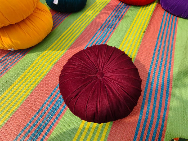 May include: A round, burgundy velvet cushion with a central button and radial stitching. The cushion is on a colorful striped mat with shades of pink, green, yellow, and blue. Other cushions in yellow, green, and red are visible in the background.