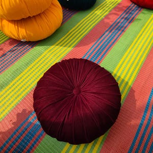 May include: A close-up shot of a round, burgundy velvet cushion with a central button and radial stitching. Other colorful round cushions are visible in the background, resting on a striped mat with green, yellow, blue, and pink stripes.