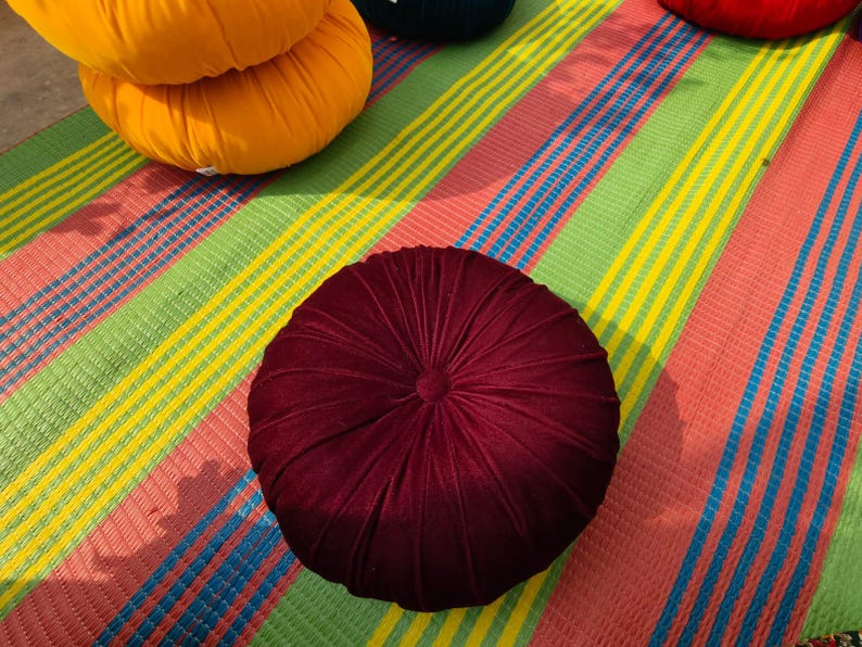 May include: A close-up shot of a round, burgundy velvet cushion with a central button and radial stitching. Other colorful round cushions are visible in the background, resting on a striped mat with green, yellow, blue, and pink stripes.