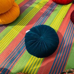 May include: A collection of colorful, round velvet cushions in shades of teal, orange, red, and burgundy. The cushions are arranged on a striped mat with a pattern of red, green, yellow, and blue stripes. The cushions have a button in the center.