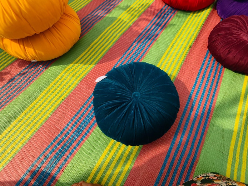 May include: A collection of colorful, round velvet cushions in shades of teal, orange, red, and burgundy. The cushions are arranged on a striped mat with a pattern of red, green, yellow, and blue stripes. The cushions have a button in the center.