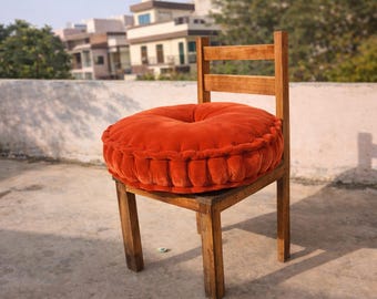 Op de afbeelding: Een houten stoel met een rond, oranje kussen. Het kussen heeft een gecapitonneerd ontwerp en is op de zitting van de stoel geplaatst. De stoel is gemaakt van lichtbruin hout en bevindt zich buiten.