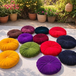 May include: A collection of colorful, round floor cushions in various shades, including yellow, purple, green, red, and blue. The cushions are arranged on a light-colored rug, with plants and flowers in the background.