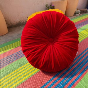 May include: A round, red cushion with a pleated design. The cushion is placed on a colorful striped mat with green, yellow, pink, and blue stripes. The cushion appears to be made of a soft fabric.