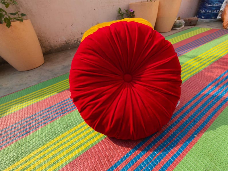 May include: A round, red cushion with a pleated design. The cushion is placed on a colorful striped mat with green, yellow, pink, and blue stripes. The cushion appears to be made of a soft fabric.