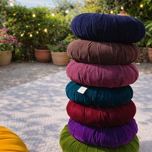 May include: A stack of seven round, velvet meditation cushions in various colors: navy, brown, burgundy, teal, red, purple, and green. The cushions are arranged outdoors with plants and string lights in the background.