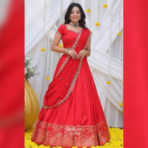 May include: A woman in a striking red lehenga choli, a traditional Indian garment. The outfit includes a short-sleeved top and a flowing skirt with gold embroidery. The ensemble is completed with jewellery and a matching red dupatta.