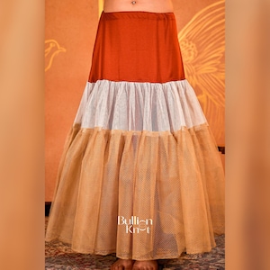 May include: A layered skirt featuring a rust-coloured top section, a white middle panel, and a tan mesh lower panel. The skirt is suitable for dance or performance. The brand name "Bullion Khit" is displayed on the skirt.