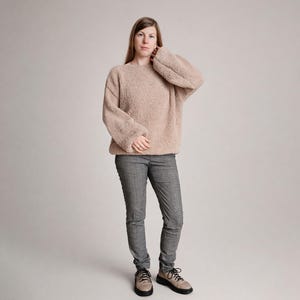 Puede incluir: Una mujer con un suéter holgado marrón claro de manga larga y pantalones de cuadros grises. El suéter tiene una apariencia texturizada. También lleva zapatos beige con cordones negros.