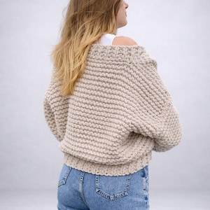 May include: A beige, chunky knit sweater with a wide neckline and long sleeves. The sweater is paired with light blue denim jeans. The model is viewed from the back, with long blonde hair.