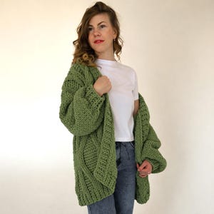 May include: A green, chunky knit cardigan with a cable knit pattern. The long-sleeved sweater is worn over a white t-shirt and blue jeans. The cardigan is made of a soft, thick yarn, perfect for cooler weather.