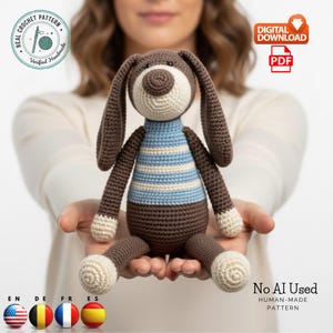 May include: A handmade crochet dog toy with brown, white, and blue accents. The dog has long floppy ears, a striped shirt, and a round nose. The image includes text that says "Real Crochet Pattern" and "No AI Used, Human-Made Pattern."