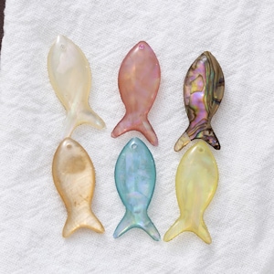 May include: Six iridescent fish-shaped pendants in various colors: cream, pink, abalone, gold, blue, and yellow. Each pendant has a small hole at the top for stringing. The pendants are arranged on a white, textured surface.