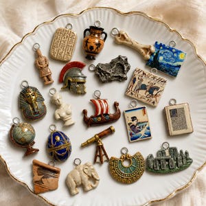 Può includere: Una collezione di vari ciondoli a tema storico e culturale, tra cui un mappamondo, un elmo romano, una nave vichinga, un busto egizio, un telescopio, un mammut, Stonehenge, un osso e ciondoli rettangolari ispirati all'arte con anelli.