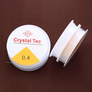 May include: Two spools of clear, elastic beading cord. One spool is round with the text "Crystal Tec" and "0.4" on the label. The other spool is rectangular. The background is a solid brown.