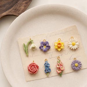 May include: A collection of eight small, three-dimensional floral charms featuring a snowdrop, purple pansy, yellow daffodil, white daisy, pink rose, blue hyacinth, pink gladiolus, and purple aster, each with a small gold-toned metal jump ring.