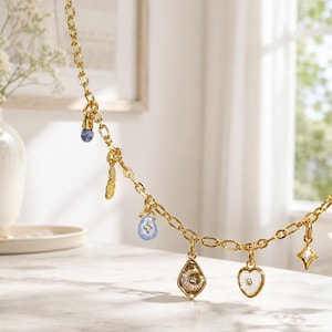 Puede incluir: Un collar de cadena dorada con múltiples dijes colgantes, incluyendo una piedra azul, una pluma dorada, una gota azul claro, un dije en forma de diamante con una luna creciente y estrella, un corazón blanco y una estrella de cuatro puntas.