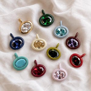 Puede incluir: Una colección de doce dijes ovalados, cada uno con una piedra preciosa facetada engastada en un marco de metal de color. Los dijes vienen en varios colores, incluyendo oro, verde oscuro, azul claro, azul marino, crema, oliva, morado, turquesa, rojo, burdeos y rosa pálido.
