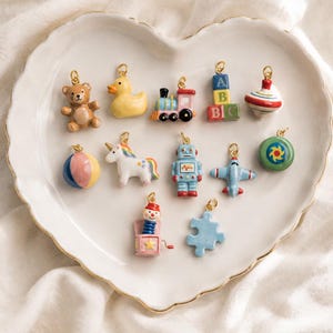 Puede incluir: Una colección de doce pequeños dijes de metal y esmalte con forma de juguetes, que incluyen un oso de peluche marrón, un pato amarillo, un tren, bloques ABC, una peonza, una pelota, un unicornio, un robot, un avión, un disco verde, una caja sorpresa y una pieza de rompecabezas azul.