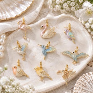 Könnte beinhalten: Eine Sammlung von neun goldfarbenen Metallanhängern mit Emaille-Vogelmotiven, darunter ein Flamingo, Schwan, Papagei, Reiher, Kolibri, eine Möwe, ein Rotkehlchen, ein gelber Singvogel und ein Tukan, jeweils mit einem kleinen Goldring.
