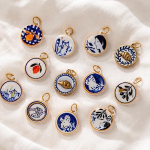 May include: A collection of eleven circular ceramic charms featuring blue and white hand-painted designs, including motifs of lemons, cats, faces, croissants, birds, coffee pots, fish, and olives. Each charm has a gold-toned metal jump ring.