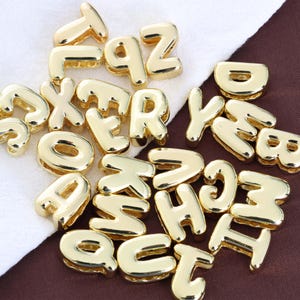 Może przedstawiać: Kolekcja błyszczących, złotych zawieszek z literami alfabetu. Litery są w pogrubionej, zaokrąglonej czcionce i są rozrzucone na białej i brązowej powierzchni. Zawieszki są prawdopodobnie używane do tworzenia biżuterii lub projektów rzemieślniczych.