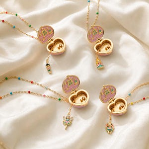 May include: Four gold-tone heart-shaped locket necklaces with pink enamel interiors. Each locket features a small dangling charm of a character, including a fairy and a rabbit. The chains are accented with small, colorful enamel beads throughout.