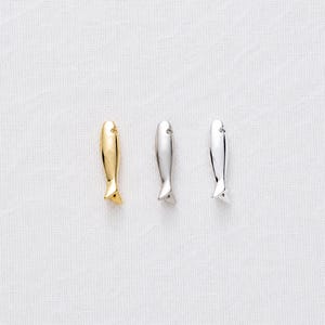 May include: Three fish-shaped pendants in gold and silver. The pendants have a smooth, polished finish and a small hole at the top. The fish have a curved body and a small tail fin. The pendants are displayed on a white background.