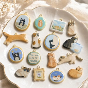 May include: A collection of small, ceramic-style cat charms featuring various feline illustrations, shapes, and colors. Several charms are circular, square, or shaped like cats, with one round charm displaying the text 'You are made of Magic.'