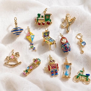 Puede incluir: Una colección de dijes de metal en tono dorado con diversas formas, incluyendo un tren, un trompo, una cometa, una máquina de pinball, una muñeca rusa, un caballito de balancín, un robot, un reloj de arena, una bicicleta y una resortera con esmalte.