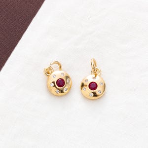 May include: A pair of gold circular charms, each featuring a central round red gemstone surrounded by six small clear accent stones. Each charm includes a small gold jump ring attached to the top for hanging, showcasing a polished finish.