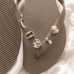 May include: A pair of taupe flip-flops featuring textured straps adorned with various metallic and pearl-like charms. The embellishments include a small silver chain with pearls, a gold rectangular frame, a gold seashell, and a round faceted stone.